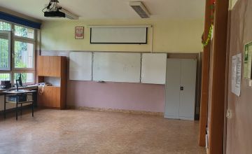 Sala szkolna bez ławek. ściany w kolorze brudnego różu. Na ścianie wisi tablica. Z lewej strony stoi biurko.